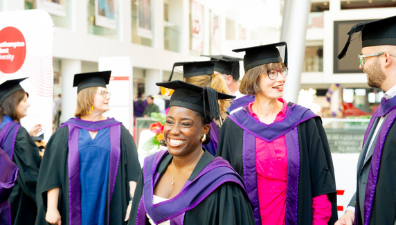 A group of graduates in caps and gowns smile and chat together at a graduation ceremony in a bright indoor venue.