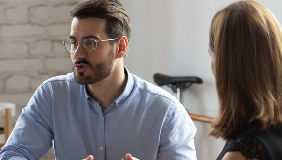 Man in glasses speaks during a meeting, gesturing as a colleague listens, with soft blue graphic shapes overlaid.