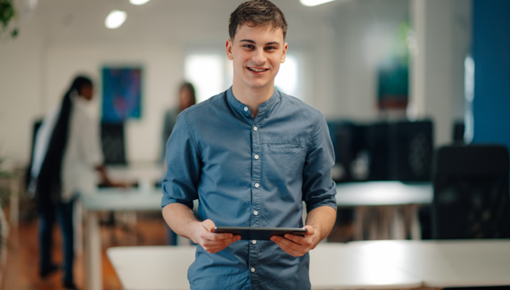 Young man stands in a modern office holding a tablet and smiling, with blurred colleagues in the background and purple graphic shapes overlaid.