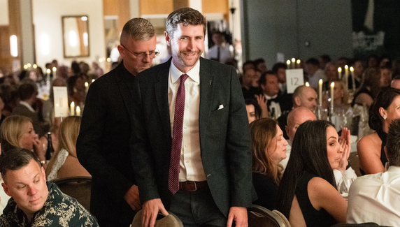 Man in a suit walks through a seated audience at a formal awards dinner, with guests at candlelit tables applauding around him.