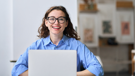 Smiling woman with glasses sits at a desk using a laptop, in a bright workspace with soft blue graphic shapes overlaid.