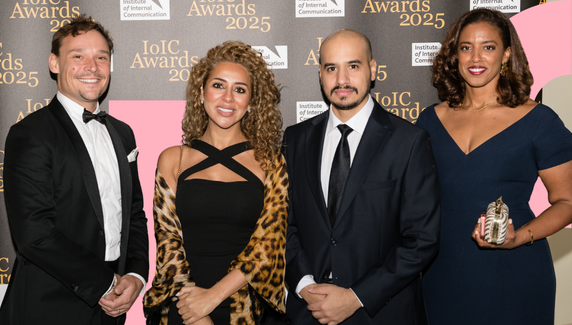 Four people dressed in formal evening wear pose at the IoIC Awards 2025, smiling in front of a branded backdrop, one holding an award.