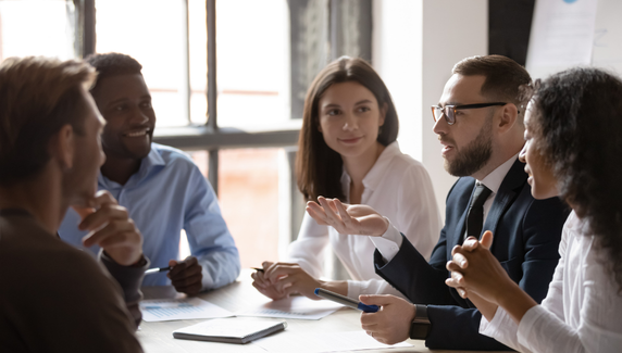 Team in a meeting around a table, discussing ideas with notebooks and a phone visible, one man gesturing as he speaks while others listen.