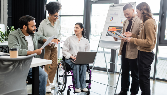 Colleagues reviewing charts and documents in a bright office, with one team member in a wheelchair leading discussion beside a presentation board.