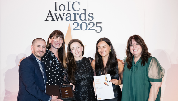 Five people smiling on stage at the IoIC Awards 2025, holding a trophy and certificate in front of a branded backdrop.