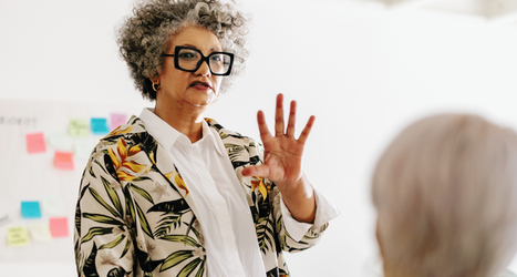 Senior woman with curly grey hair and glasses speaking and gesturing in a meeting, with blue graphic shapes overlaid.