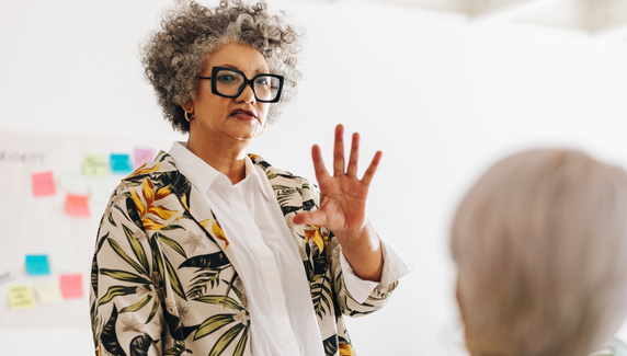 Senior woman with curly grey hair and glasses speaking and gesturing in a meeting, with blue graphic shapes overlaid.