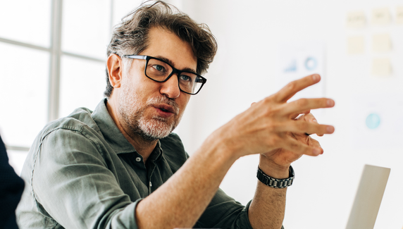 Man with glasses and grey hair gestures while speaking in a meeting, seated beside a laptop against a bright, modern office background.